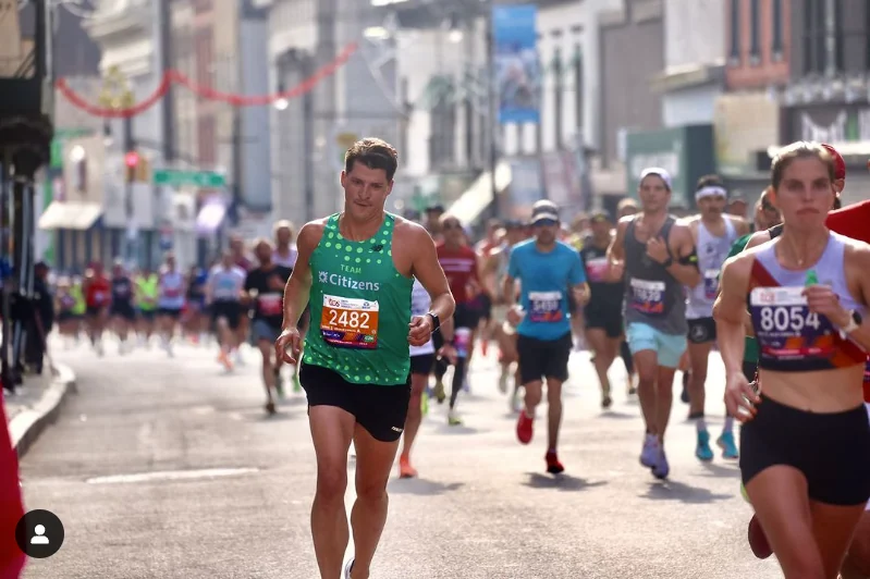 A runner in the green Team Citizens singlet with its halftone dot pattern, bib number 2482, running down a Brooklyn street mid-race surrounded by other marathon runners.