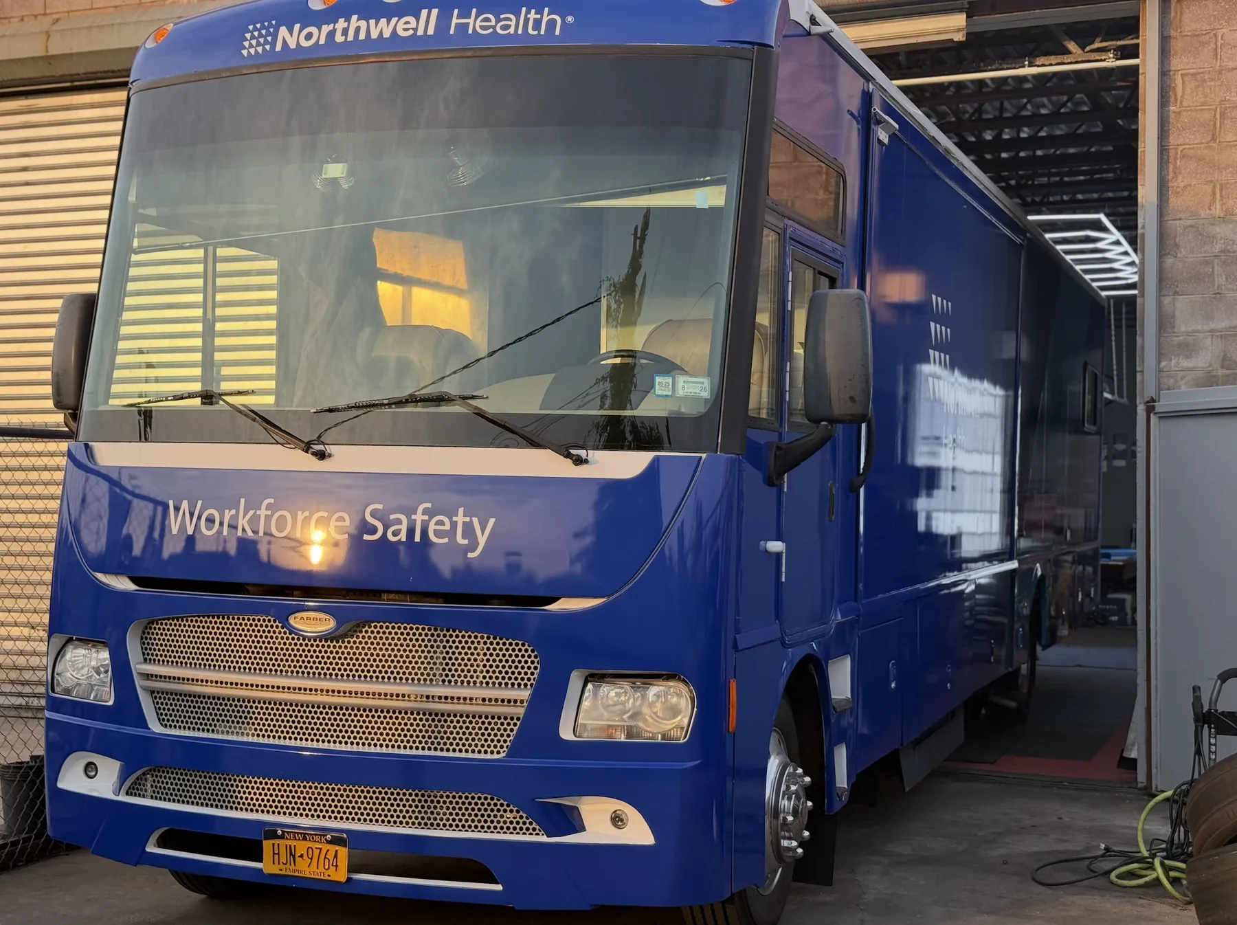 Photograph of the finished wrapped RV in a garage bay at dusk, deep blue with the Northwell Health mark above the windscreen and Workforce Safety across the nose.