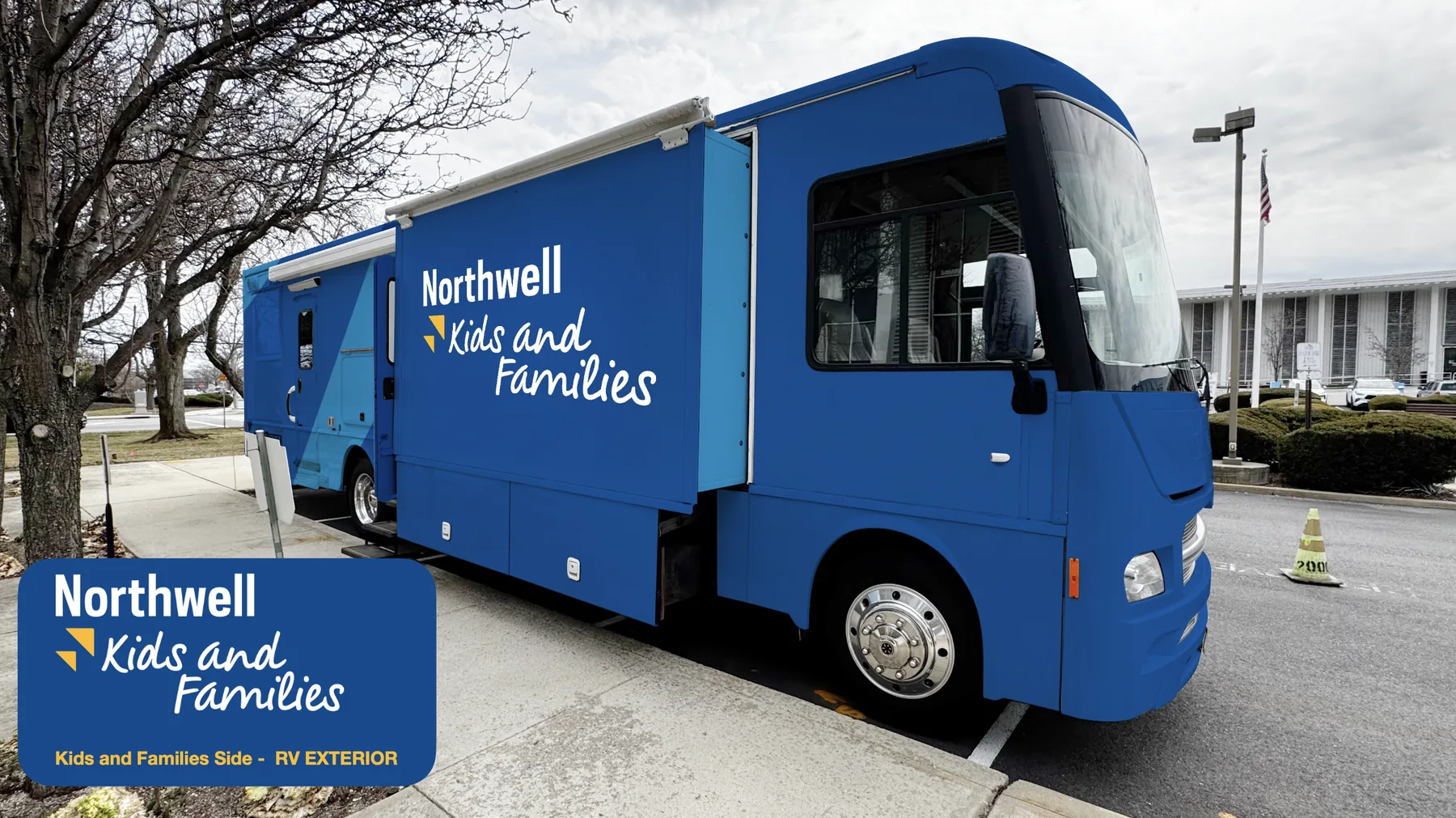 Exterior wrap rendering, passenger side: the RV parked under bare trees with the slide-out extended, wrapped in two blues with the diagonal split running through the slide-out and the Kids and Families logo forward on the body.