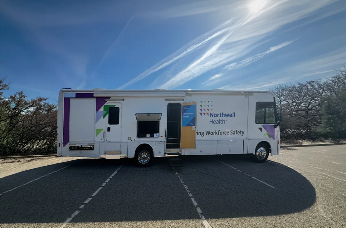 The RV before the rebuild: a white recreational vehicle in an empty car park, carrying the Northwell Health mark, a Workforce Safety line and small purple and green chevrons, with its door open and a Welcome sign propped inside.