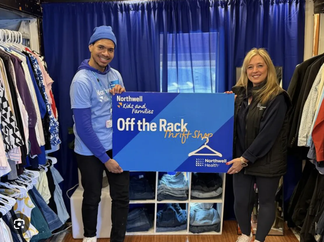 Two team members inside the stocked shop holding the Off the Rack Thrift Shop board, surrounded by hanging clothes and folded jeans in cube shelving.