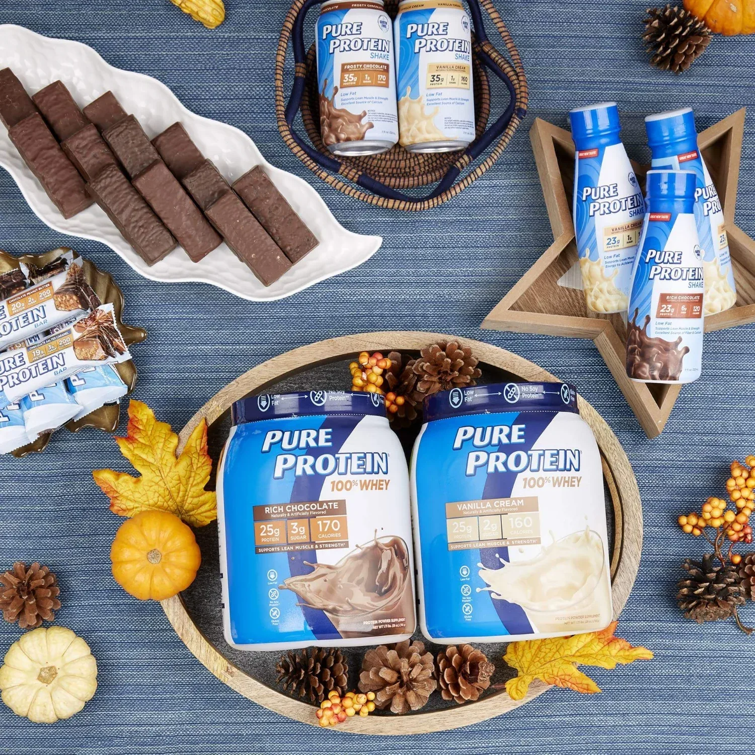 Autumn flat lay on denim with pumpkins, pinecones and maple leaves surrounding Pure Protein powders, shakes and bars.