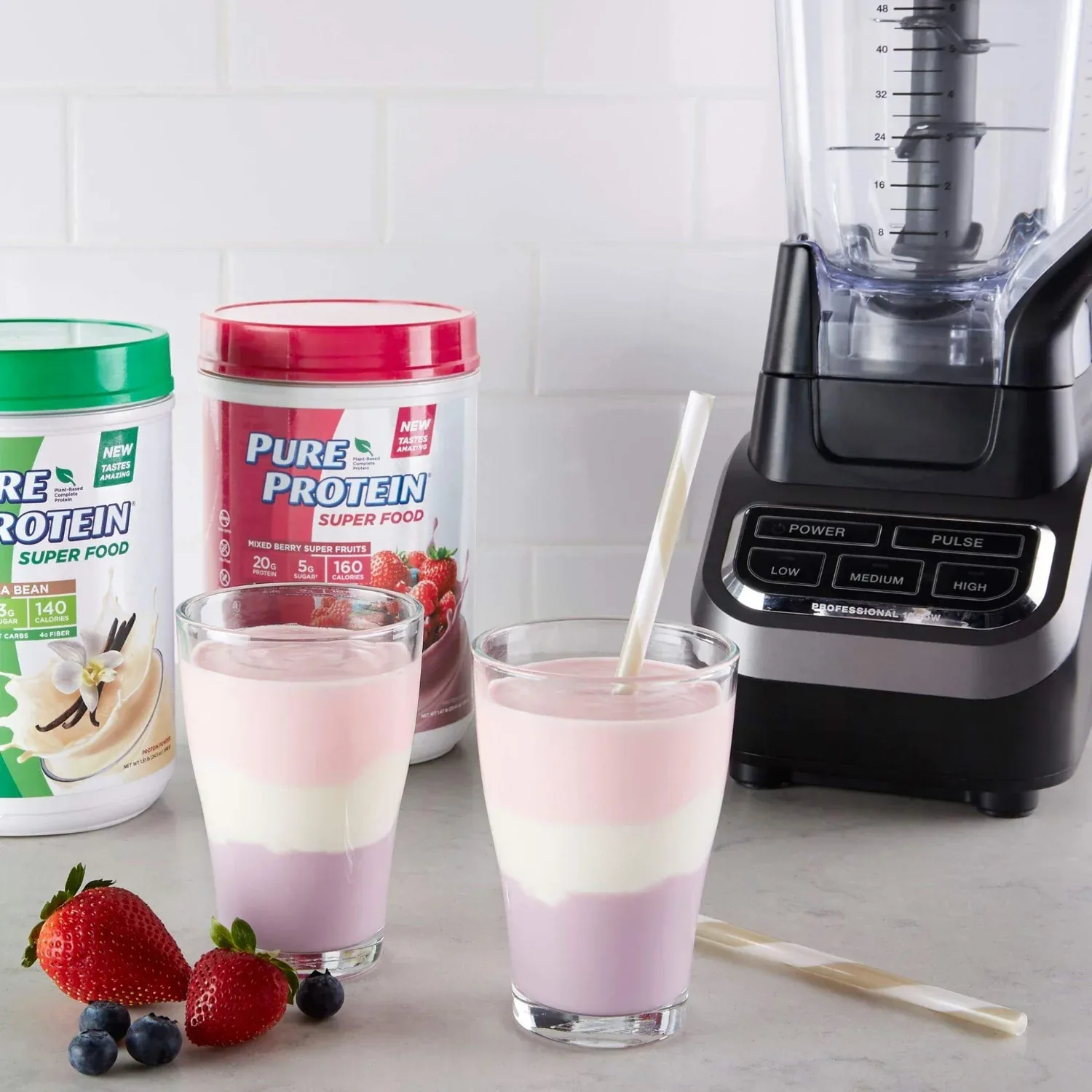 Two glasses of tri-colour layered smoothie beside Pure Protein Super Food tubs, a blender and fresh berries.
