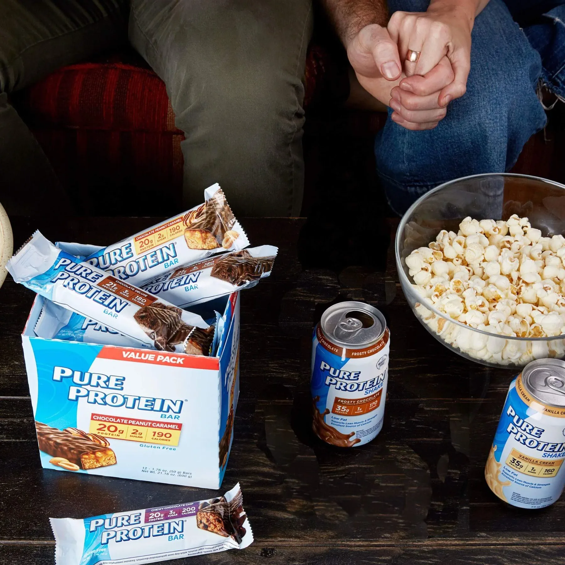 Two people seated on a sofa with popcorn, Pure Protein bars and shake cans on a dark wooden coffee table.