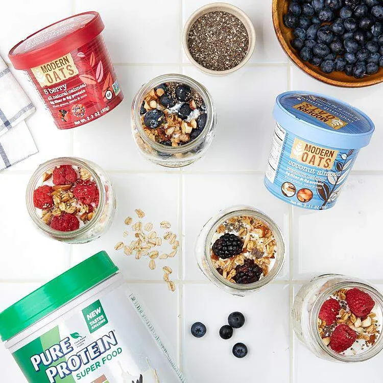 Overnight oats in glass jars topped with berries and nuts, arranged on white tile with Modern Oats cups and a Pure Protein Super Food tub.