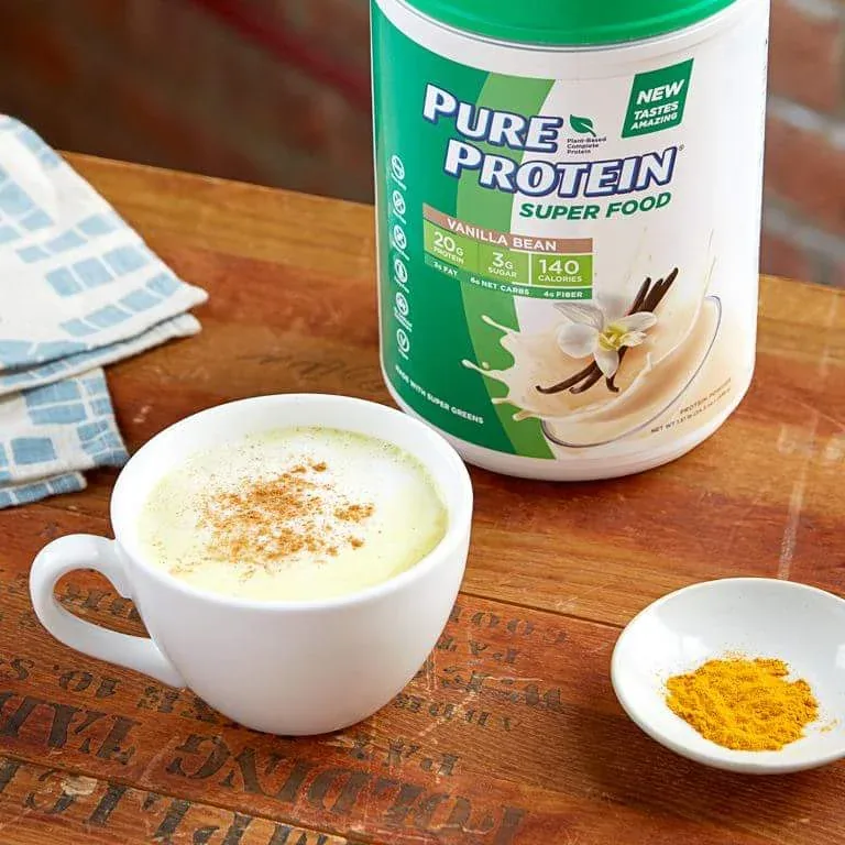 A golden turmeric latte dusted with spice on a worn wooden table beside a Pure Protein Super Food Vanilla Bean tub.