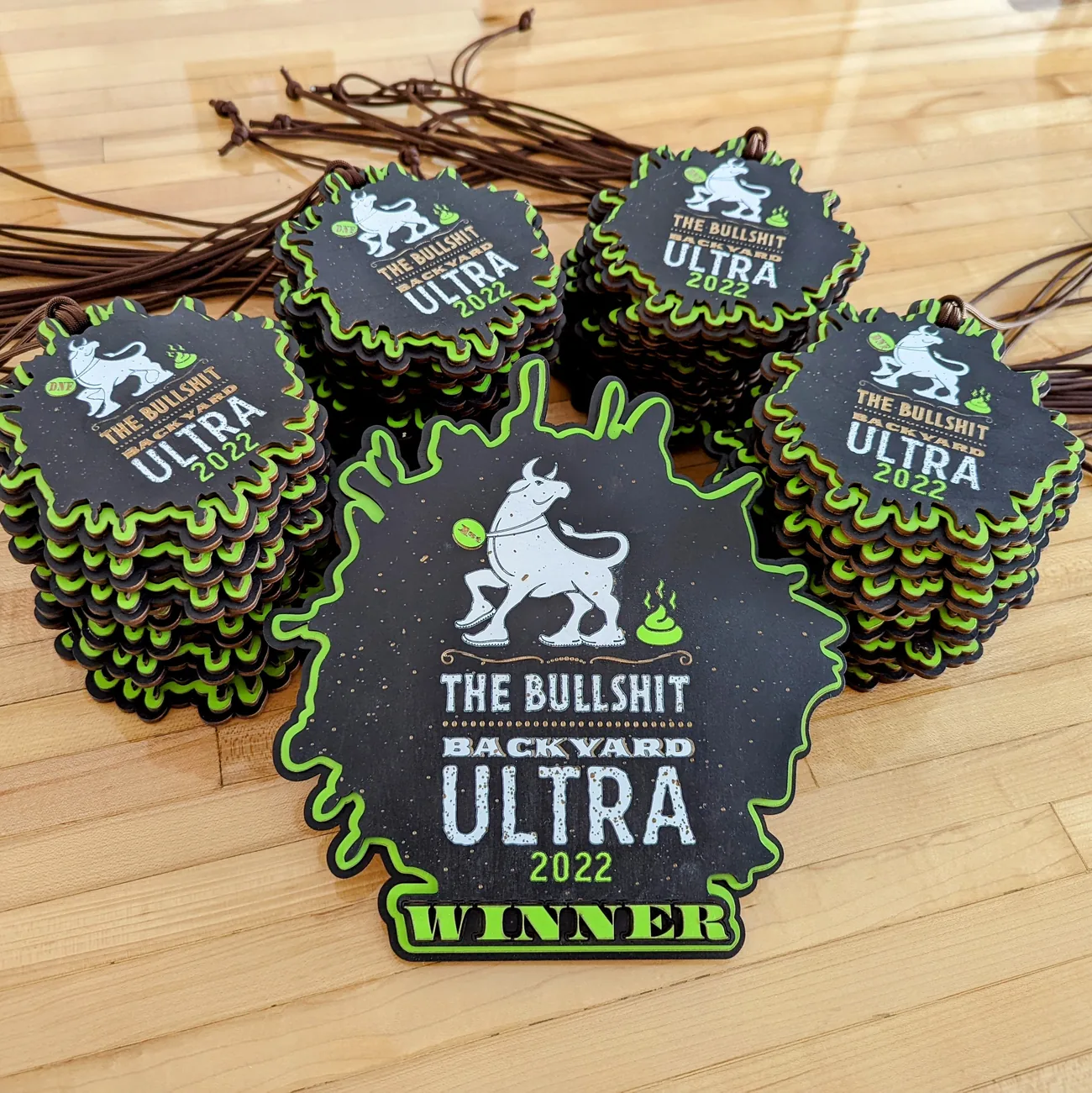 Stacks of black and lime green Bullshit Backyard Ultra 2022 awards on a wooden floor with brown cords, each shaped like a splat and showing a cartoon bull in running shoes, with the larger front piece reading WINNER and the stacked ones marked DNF.