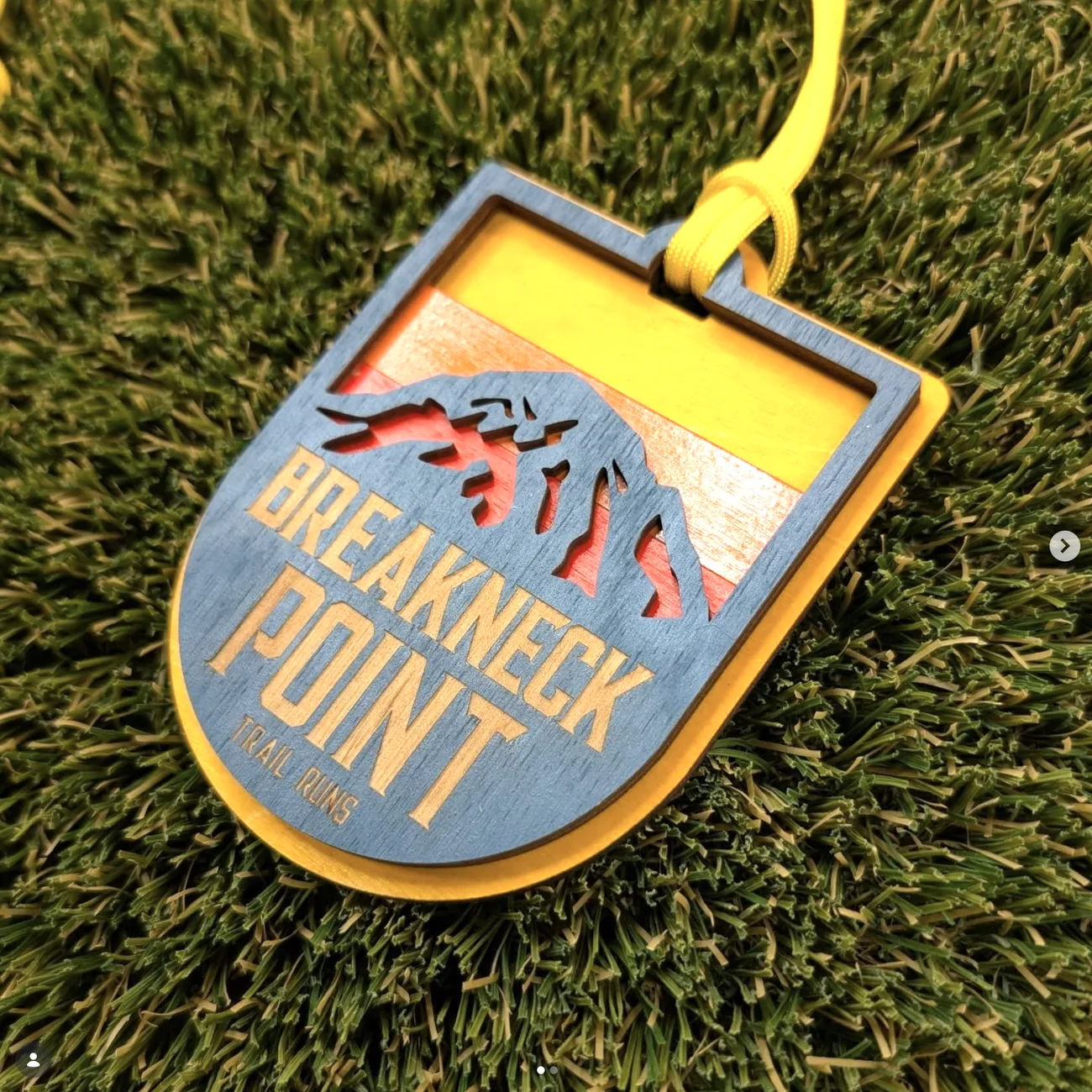 Breakneck Point Trail Runs medal on turf: a rounded blue-stained shield over a yellow base layer, with a mountain ridge and a leaping runner cut out to reveal an orange and cream sunset behind, the event name engraved into the blue face.