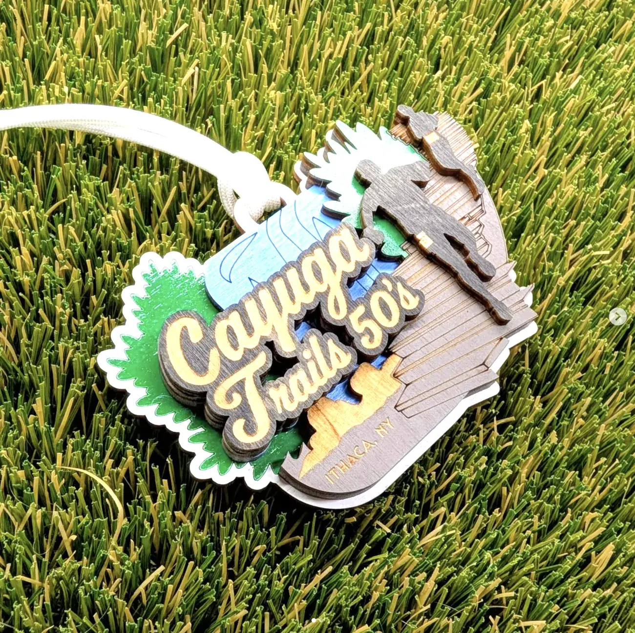 Cayuga Trails 50s medal on turf, layered in green, blue and stained grey, with a waterfall and gorge cut into the right side, a runner silhouette on the clifftop, script event lettering and Ithaca NY engraved at the base.