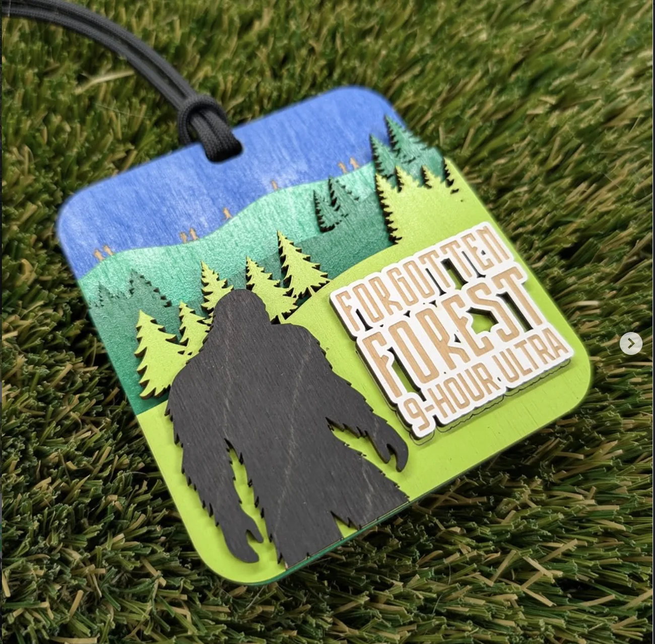 Square Forgotten Forest 9-Hour Ultra medal on turf, with layered bands of blue sky, teal and lime green hills, cut pine tree silhouettes and a dark bigfoot figure in the foreground, and the event name in a white and wood-toned block at right.