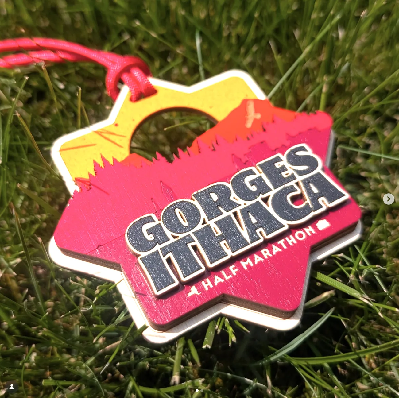 Star-shaped Gorges Ithaca Half Marathon medal on grass in yellow, orange and hot pink stained layers, with a cut treeline silhouette, a circular void for the sun and the event name in dark block letters.