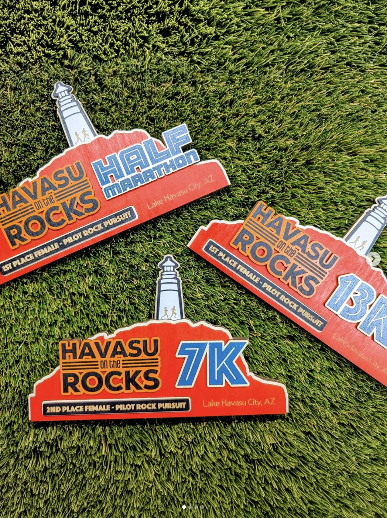 Three Havasu on the Rocks awards on turf in red stained ply with a lighthouse rising from a rocky ridge, each engraved with a different distance — Half Marathon, 13K and 7K — and a placing band beneath the event logo.
