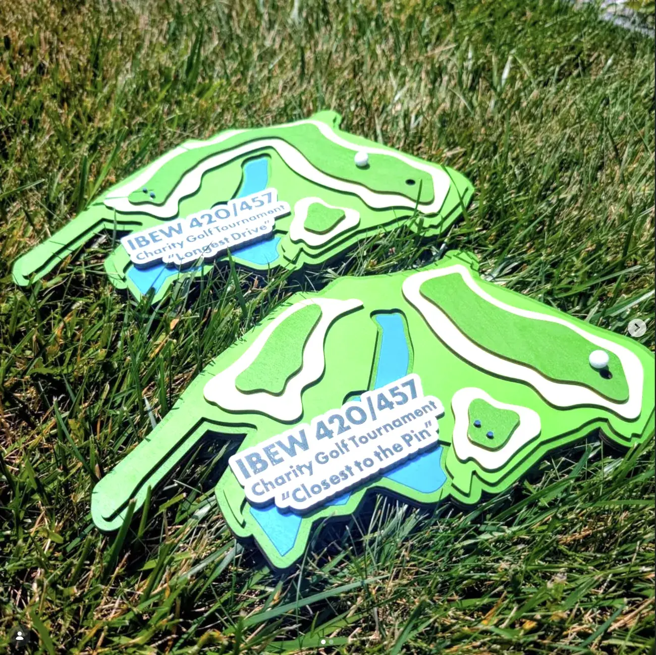 Two IBEW 420/457 Charity Golf Tournament awards lying in grass, each cut to the plan shape of a golf hole in layered greens with white sand bunkers, a blue water hazard and a small white ball marking the pin, engraved Longest Drive and Closest to the Pin.