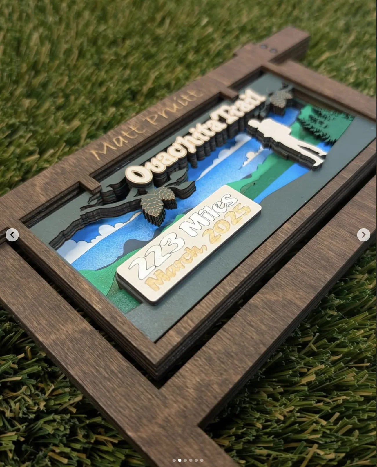 The Ouachita Trail trophy photographed at an angle on turf, showing the depth of the layered scene inside the frame — clouds, ridgelines, pine cones, a hiker silhouette and a raised plaque — with the recipient name engraved along the top rail.