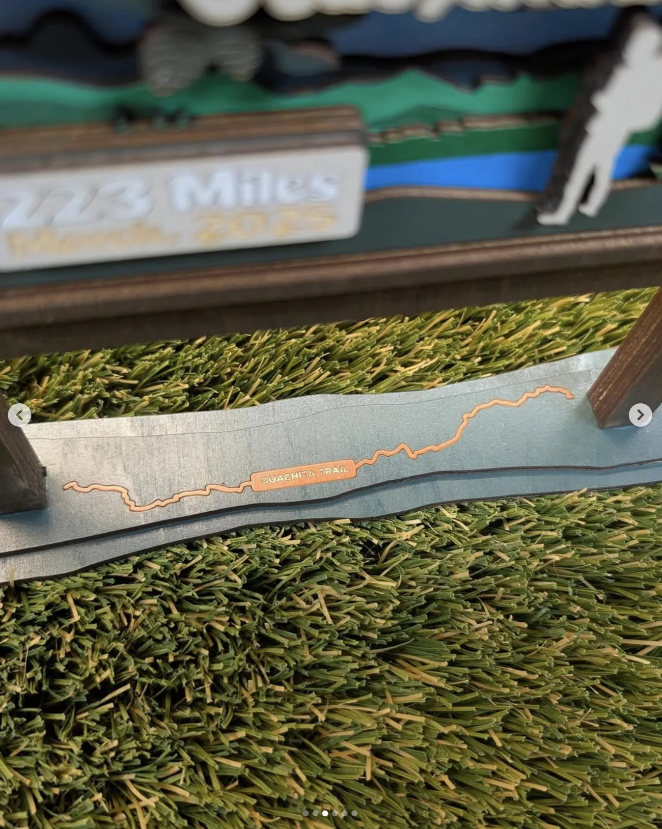 Close detail of the trophy's base, a stained green plank cut to a wave profile with the winding route of the Ouachita Trail inlaid in copper-toned wood and labelled along its length.