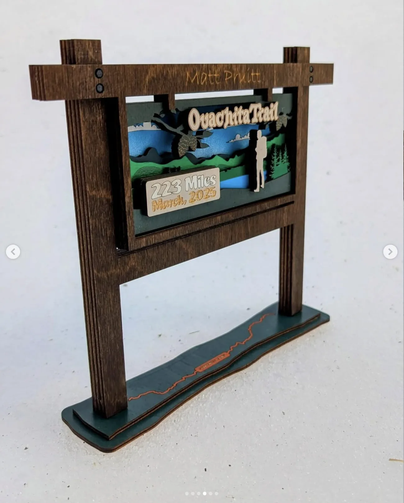 Freestanding wooden trophy built as a miniature trail head sign, with two posts and a crossbar carrying an engraved recipient name, framing a layered scene of mountains, pines, a hiker and a plaque reading 223 Miles, March 2025, mounted on a green base.