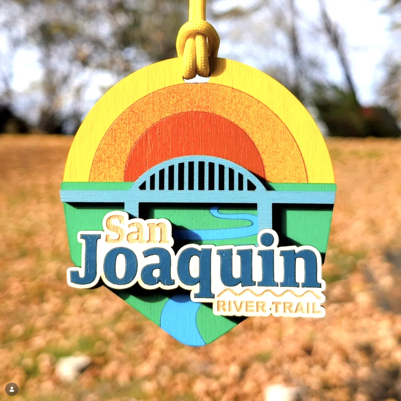 Round San Joaquin River Trail medal hanging on a yellow cord, built from concentric stained rings of yellow, orange and red forming a sunburst behind a pale blue arched bridge, with green banks and a blue river below and the event name in layered lettering.