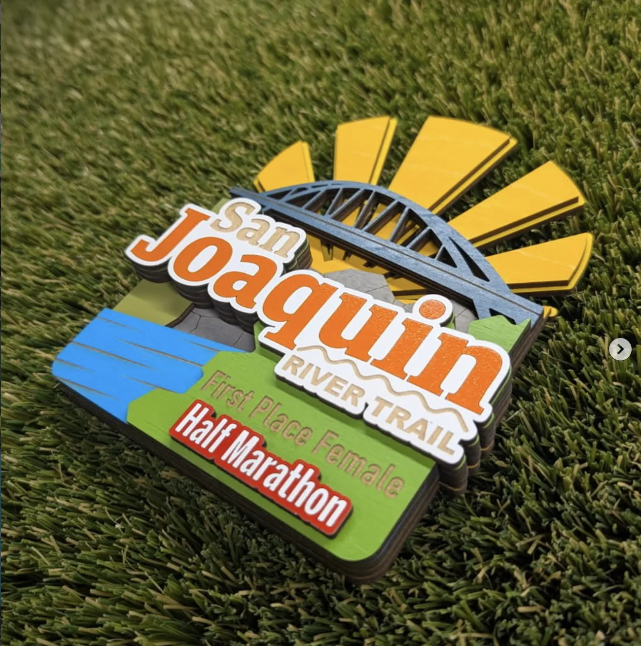 San Joaquin River Trail first place female half marathon award on turf, a layered plaque with a yellow sunburst behind a blue truss bridge, green banks, a blue river and the event name in orange and cream lettering.
