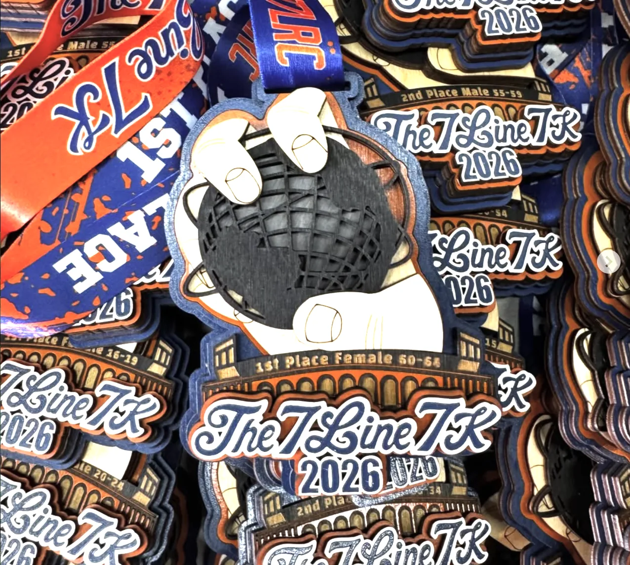 A large batch of The 7 Line 7K 2026 awards piled together, each showing a baseball glove catching a ball rendered as a globe, with blue and orange stadium arches and script lettering, and orange and blue ribbons among them.