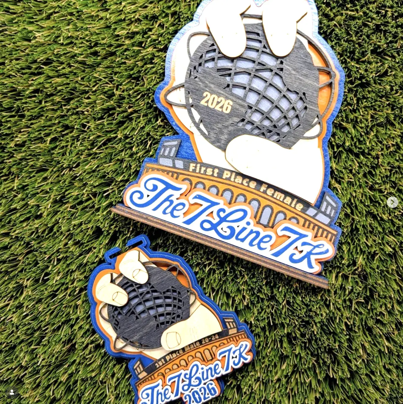 Two sizes of The 7 Line 7K award on turf, a large stand-mounted first place piece and a smaller hanging medal, both showing a glove catching a globe-patterned ball above stadium arches and script lettering.