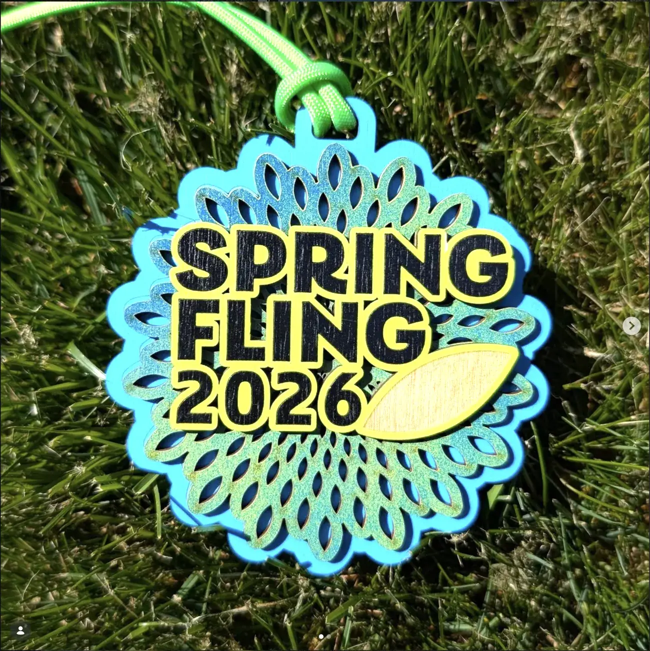 Round scalloped Spring Fling 2026 medal on grass, built as a flower with layered blue and glittered green petals, black block lettering outlined in yellow and a single yellow and wood-toned leaf at the lower right.