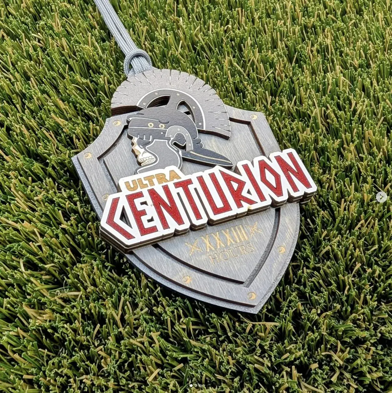 Laser-cut Ultra Centurion award: a layered wooden shield in silver-grey with a Roman centurion helmet and skull rising above it, the word CENTURION cut in red block letters across the front, and XXXIII HOURS engraved on the lower shield, resting on turf.