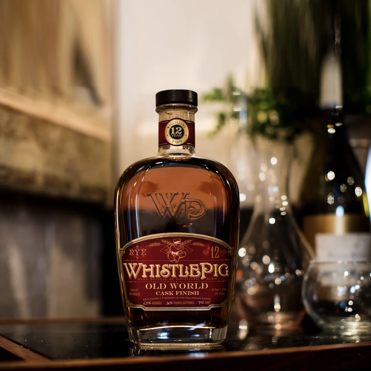 The WhistlePig 12 Year Old World Cask Finish bottle with its burgundy label, standing on a dark wooden tray with softly lit glassware and greenery blurred behind it.