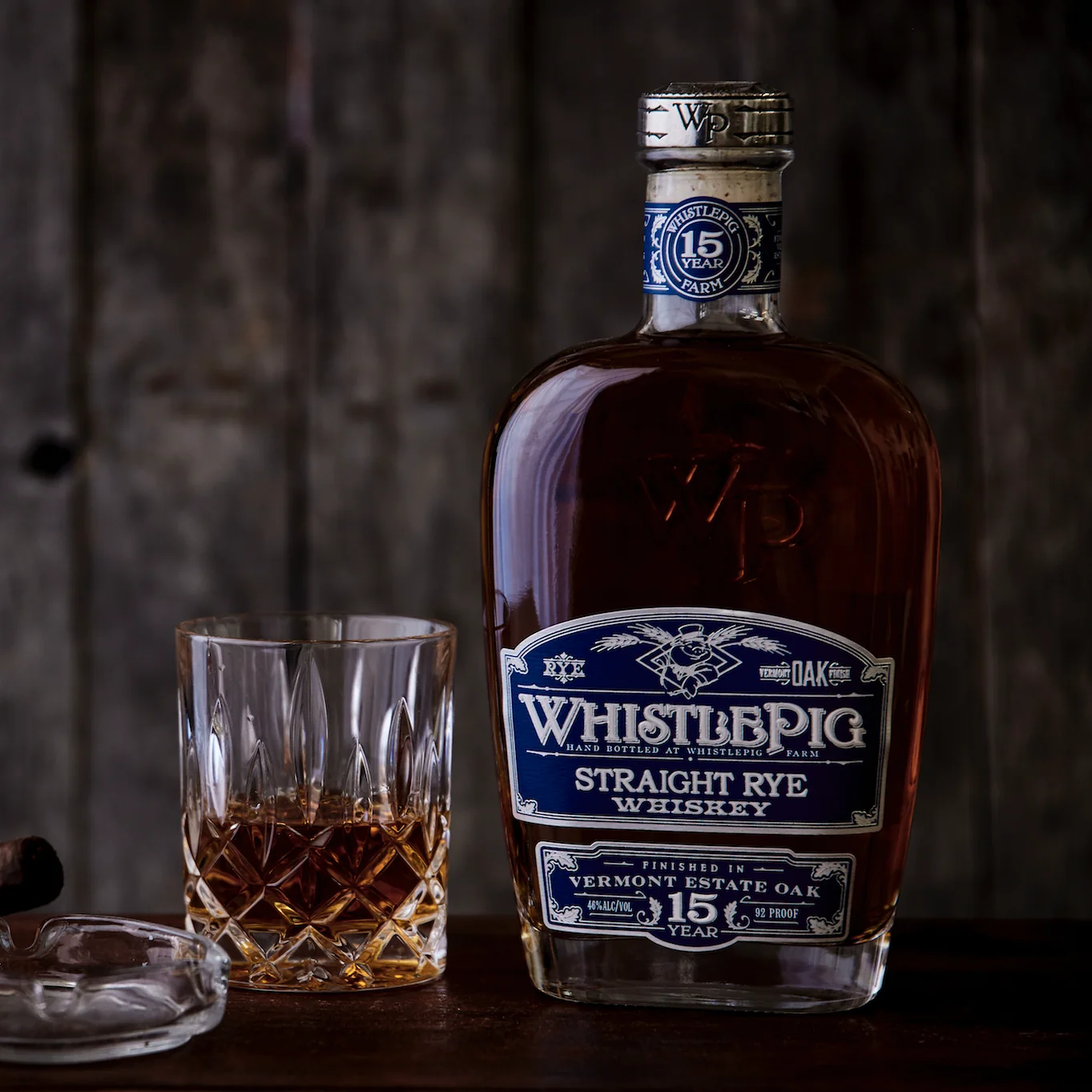 The WhistlePig 15 Year Vermont Estate Oak bottle with its deep blue label on a dark wooden surface, beside a cut crystal tumbler of whiskey, a glass ashtray and a cigar, against weathered grey barn board.