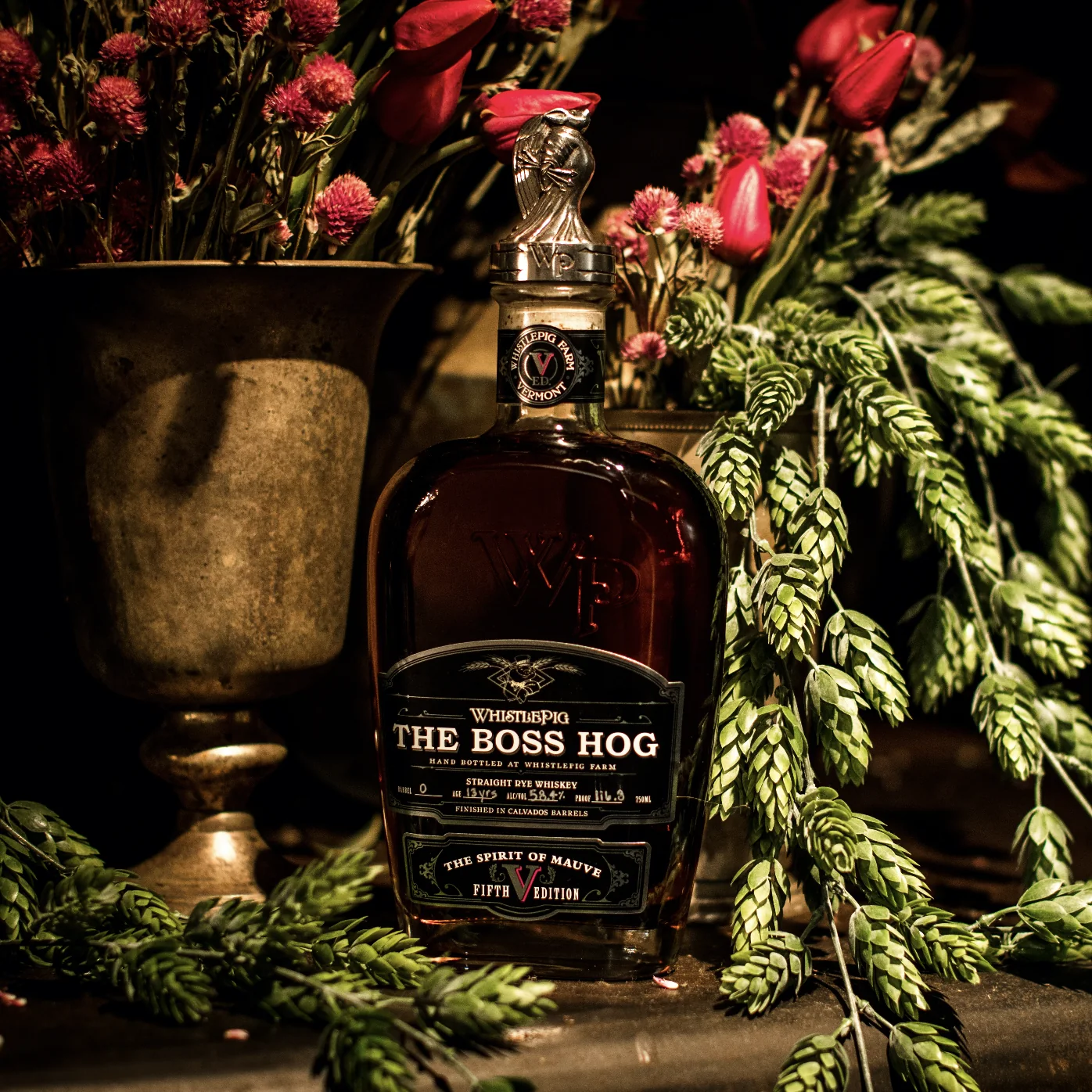 The Boss Hog Fifth Edition bottle with its silver pig stopper, lit low against dried burgundy flowers, red tulips and trailing green hop bines on a dark stone surface.