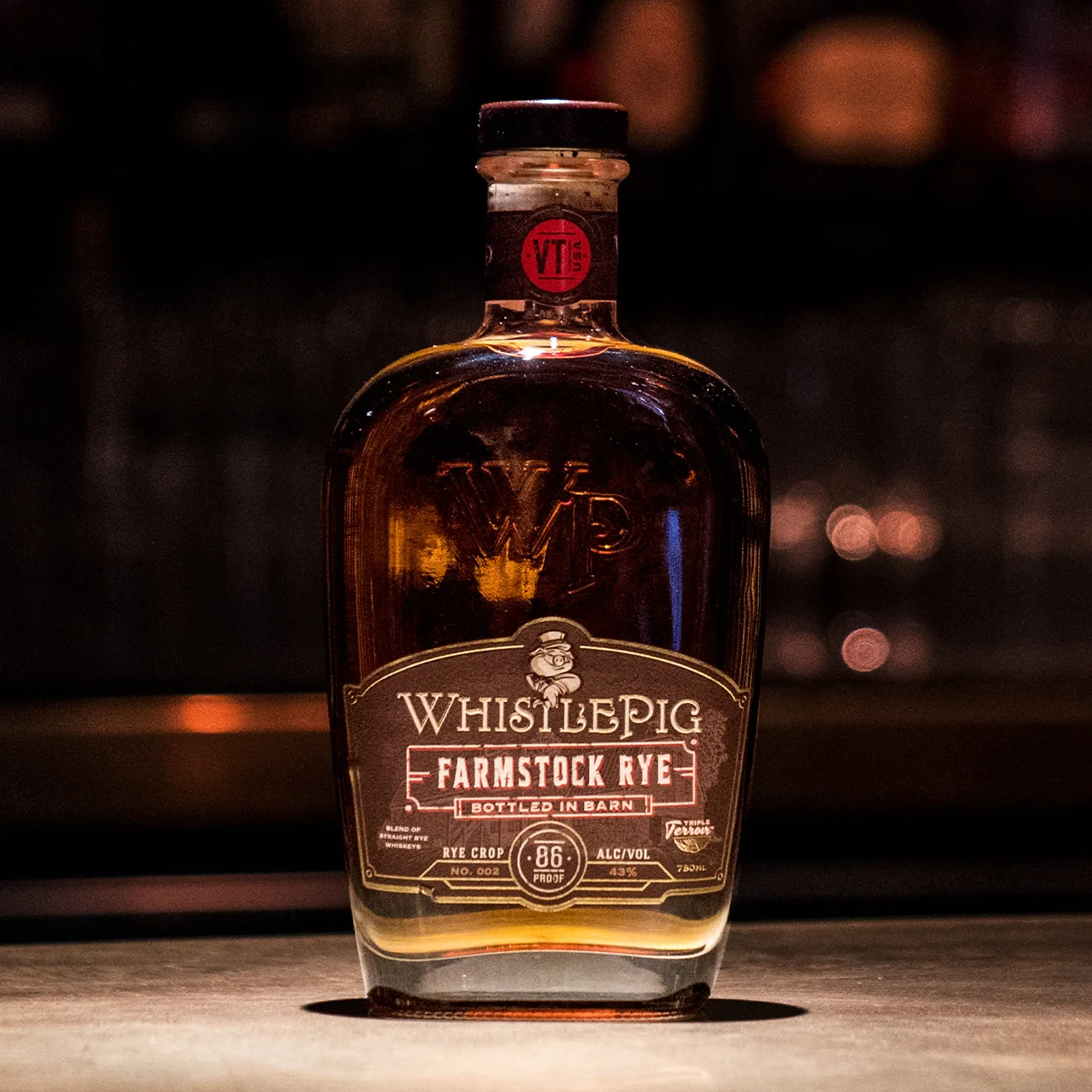 The Farmstock Rye bottle on a concrete surface in near darkness, lit from one side with barrel shapes and warm highlights barely visible in the deep background.