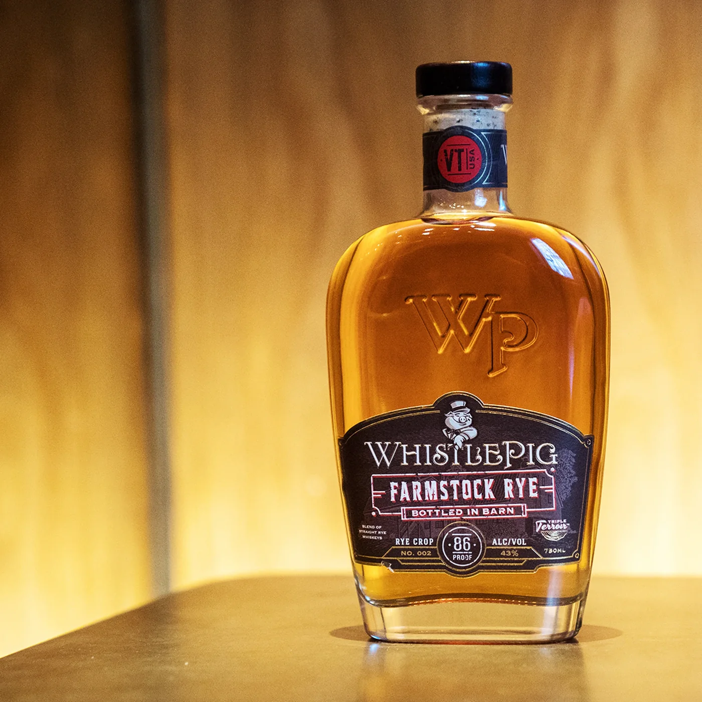 The WhistlePig Farmstock Rye bottle on a pale surface against a warm golden backdrop, lit so the whiskey glows amber and the embossed WP monogram catches the light.