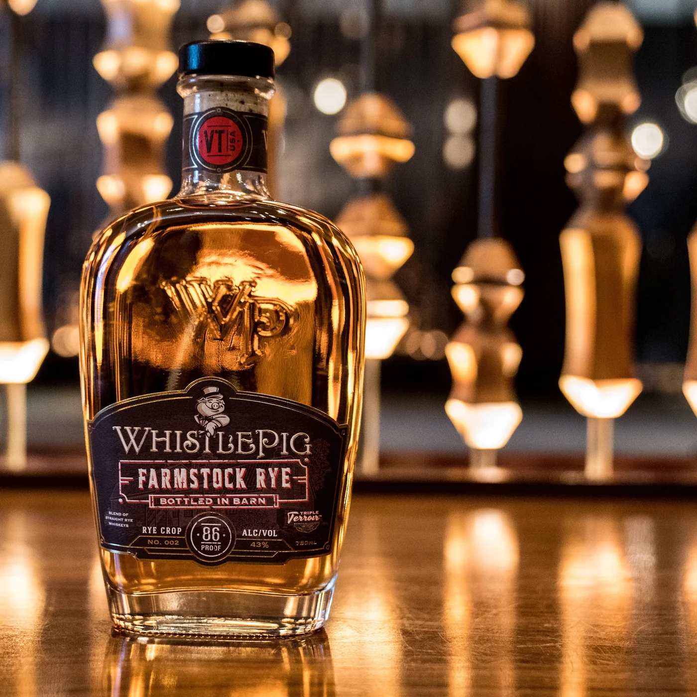 The Farmstock Rye bottle on a polished wooden bar with a row of hanging pendant bulbs thrown far out of focus behind it, their reflections running down the tabletop.