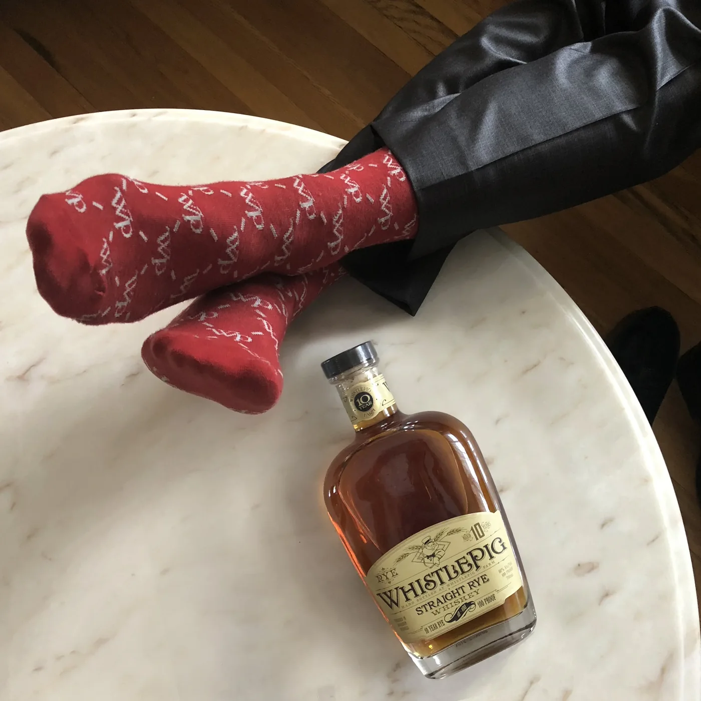 Overhead shot of crossed feet in red WhistlePig-patterned dress socks under grey suit trousers, resting on a white marble table beside a bottle of 10 Year Straight Rye.