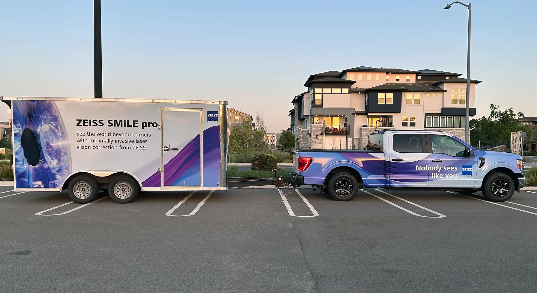 A ZEISS SMILE pro branded pickup truck and matching enclosed trailer parked at dusk, wrapped in an iridescent blue and purple corneal graphic, with the trailer reading ZEISS SMILE pro and the truck reading Nobody sees like you.